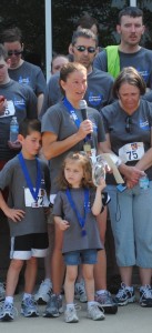 Angie Hipsher speaking at the GYRIG 5K run/walk in Indianapolis in June 2012. Her husband sands behind her and their two children.