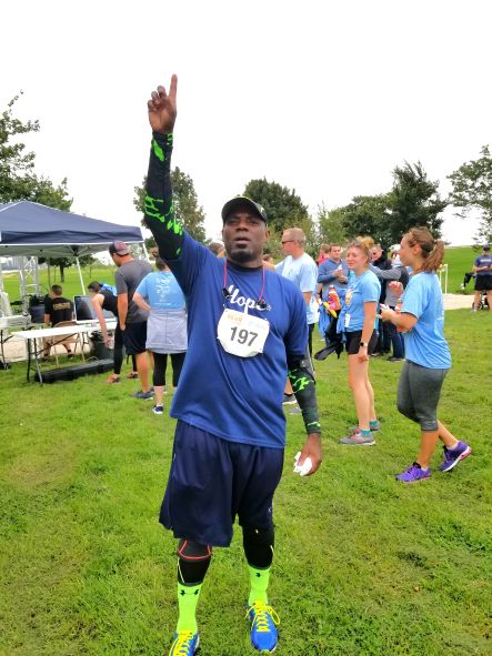 Aaron Watts, a Black man dressed in workout attire with a race bib, pointing at the sky.