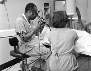 Kenneth Forde and another medical professional performing a colonoscopy. Forde is sitting on the edge of a chair with his eye up to the scope, with the patient laying on their side on the table. The photo itself is black and white 