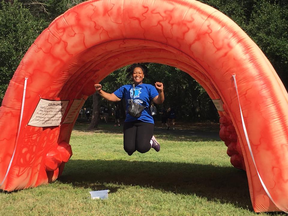Ashley Johnson, a Black woman, jumps in front of the giant, anatomically correct colon at Get Your Rear in Gear Orlando. There is a grassy field and trees in the background