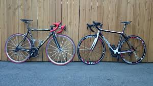 Two road bikes, one with red handle bars and tires, the other with black handle bars and tires, resting against a tall wood fence
