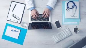 A doctor types on a Mac computer, surrounded by various items such as a clipboard, a glass of water, a thermometer, stethoscope.