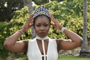 Andrea Darville is standing in front of a bush. She's wearing a white dress and a crown with blue and silver jewels. 