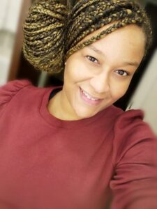 Ashley Johnson smiles at the camera. She is wearing a red shirt and her hair is in braids and pulled into a bun on the side of her head.