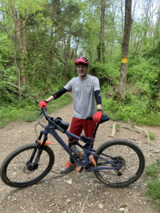 President of Olympus, Julien Sauvagnargues, on a bike. Green trees are in the background. He's wearing a gray shirt, and red shorts with a red bike helmets and gloves